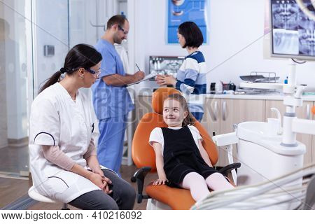 Shy Little Girl Looking Up At Dentist Doctor Waiting Teeth Cavity Treatment. Child With Her Mother D