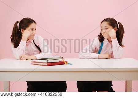 Bored Little Twin Schoolgirls Sitting Behind Schooldesk, Staring At Each Other