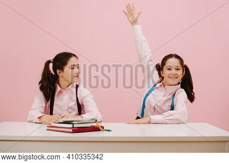 Eager Little Twin Schoolgirls Sitting Behind Schooldesk. Frontal View. Over Pink