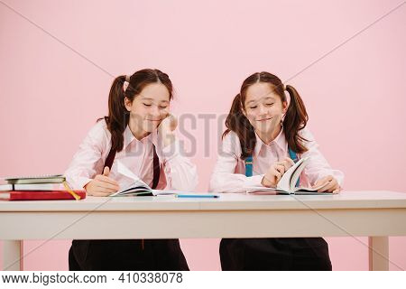Two Happy Twin Schoolgirls Sitting Behind Schooldesk. Frontal View. Over Pink