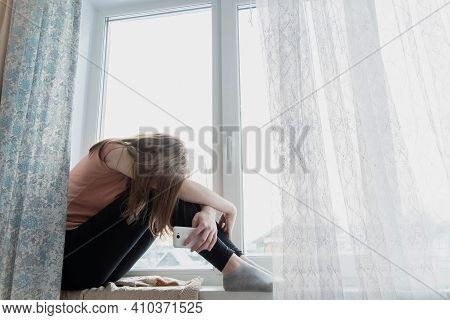 A Teenage Girl Sits On A Window Sill With Her Phone And Looks Out The Window. The Concept Of Lonelin