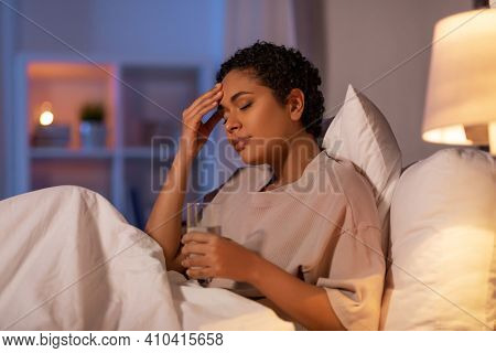 people, bedtime and rest concept - stressed african american woman having headache and drinking water in bed at night