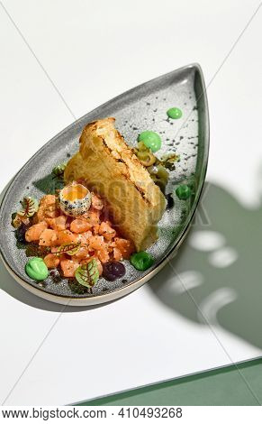 Salmon tartar with quail egg and toast bread. Raw fish tartare restaurant appetizer on white table with green wall. Day sunlight with hard shadow of monstera palm leaves. Summer or spring food concept