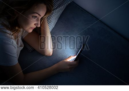 Young woman in bed holding a phone, tired and exhausted, blue light straining her eyes, messing up her circadian rhytm
