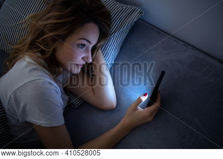 Young woman in bed holding a phone, tired and exhausted, blue light straining her eyes, messing up her circadian rhytm