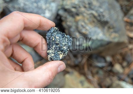 Chromite Ore In Hand, Selective Focus. Piece Of Natural Mineral Over Rocks Background