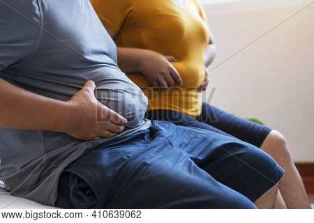 Fat Man And Woman Holding Their Big Belly While Sitting On The Bed Suffering From Extra Weight. Coup