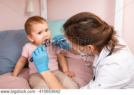 Pediatrician examines mouth of a baby girl at home during coronavirus COVID-19 quarantine. Doctor using wooden tongue depressor and torch