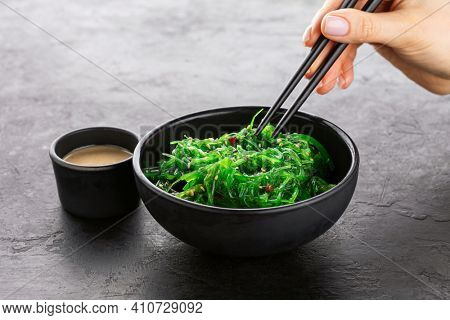 Female hand hold seaweed salad with chopsticks over black bowl on black slate background.