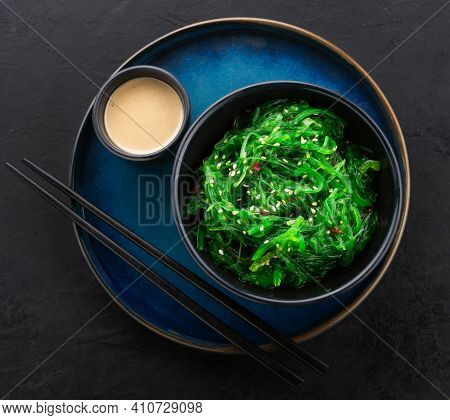 Healthy wakame seaweed salad in a black bowl, peanut sauce on side and chopsticks on a beautiful blue plate against a black slate background. View from above.