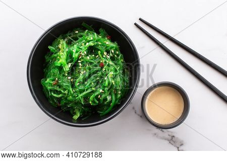 Healthy wakame seaweed salad in a black bowl, nut sauce on the side and chopsticks on a white stone background. View from above.