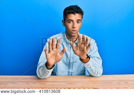 Young handsome african american man wearing casual clothes sitting on the table moving away hands palms showing refusal and denial with afraid and disgusting expression. stop and forbidden. 