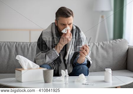 Middle-aged Bearded Man Covered In Blanket Feeling Sick, Checking For Fever With Thermometer, Sneezi