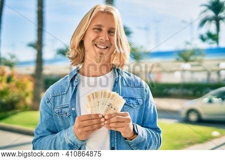 Young blond scandinavian man smiling happy holding danmark 100 kroner banknotes at the city.
