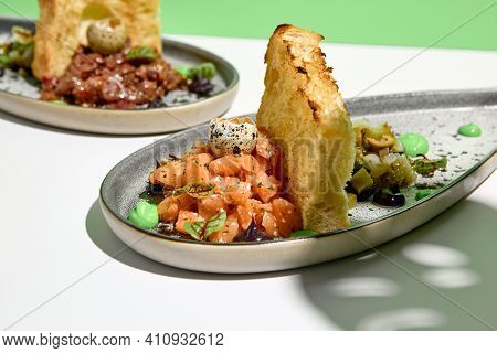Salmon tartar with quail egg and toast bread. Raw fish tartare restaurant appetizer on white table with green wall. Day sunlight with hard shadow of monstera palm leaves. Summer or spring food concept