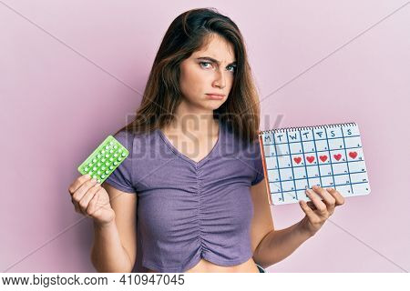 Young caucasian woman holding birth control pills and menstrual calendar skeptic and nervous, frowning upset because of problem. negative person. 