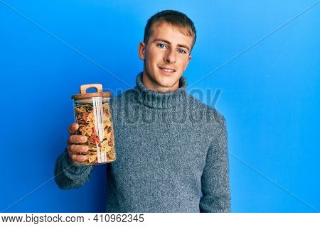 Young caucasian man holding jar of uncooked pasta looking positive and happy standing and smiling with a confident smile showing teeth 