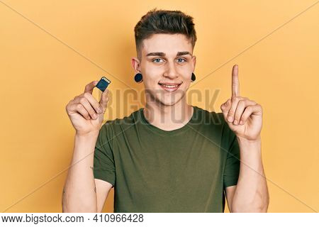 Young caucasian boy with ears dilation holding sdxc card smiling with an idea or question pointing finger with happy face, number one 