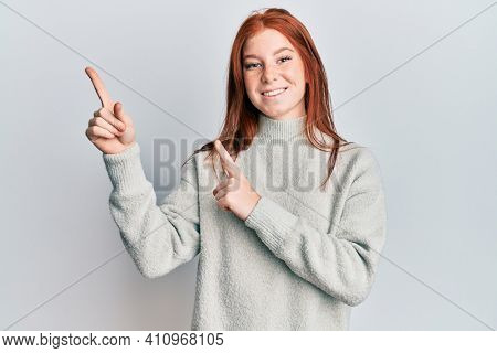 Young red head girl wearing casual winter sweater smiling and looking at the camera pointing with two hands and fingers to the side. 