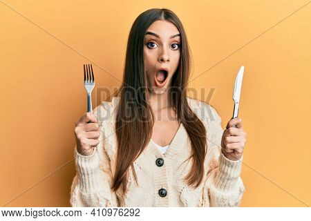 Beautiful brunette young woman holding fork and knife ready to eat afraid and shocked with surprise and amazed expression, fear and excited face. 