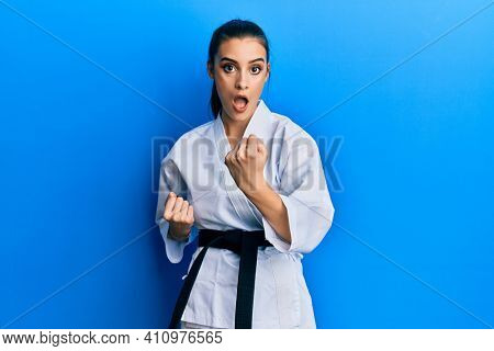 Beautiful brunette young woman wearing karate fighter uniform with black belt doing attack pose afraid and shocked with surprise and amazed expression, fear and excited face. 