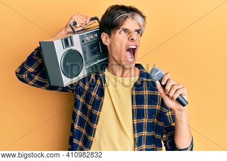 Young hispanic man singing song using microphone and boombox angry and mad screaming frustrated and furious, shouting with anger. rage and aggressive concept. 