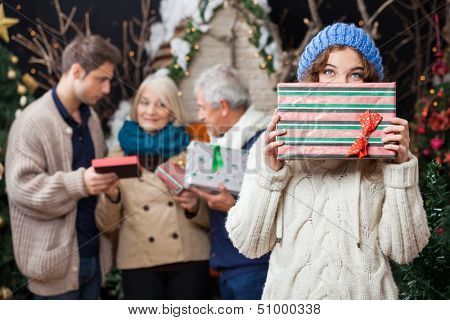 Portrait of thoughtful young woman holding Christmas present with family standing in background at store