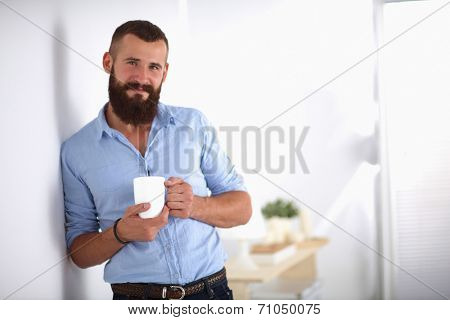 Young man  standing near wall and holding cup of coffee in offi