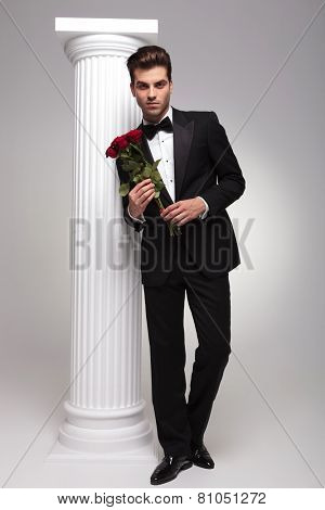 Full body picture of a elegant business man holding a bouquet of red roses in his hands while leaning on a white colomn