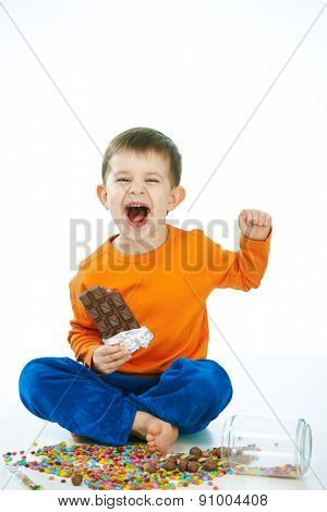 Naughty little kid eating chocolate sitting cross-legged on floor, sweets spilt. Laughing, hand in hair, isolated on white.