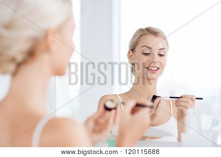 beauty, make up, cosmetics, morning and people concept - young woman applying eyeshade with makeup brush and looking to mirror at home bathroom