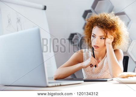 Pensive redhead curly young woman designer using laptop and grapic tablet on workplace 