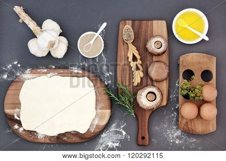 Baking ingredients with dough, yeast, eggs, olive oil, garlic, mushrooms, herbs, rye grain in lovespoon and flour dust on maple wood over grey.