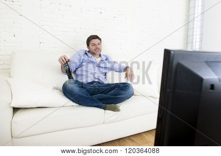 young happy man watching tv lying at home living room sofa with remote control looking relaxed smiling and laughing enjoying television program comedy movie or sitcom