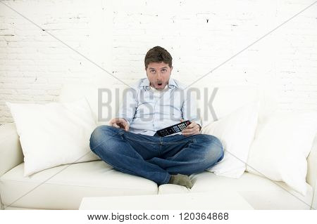 young man watching tv sitting at home living room sofa holding remote control looking intense and very interested enjoying television program or movie in disbelief amazed and shock face expression