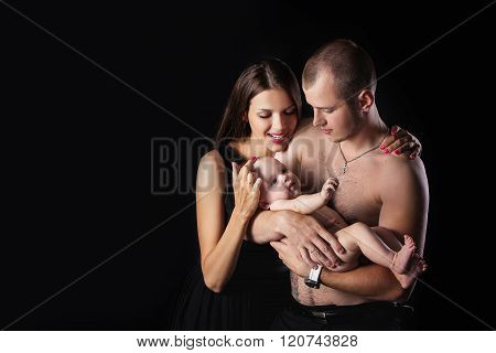 Young happy family in the studio on a dark background in dark clothes holding a baby on the hands an