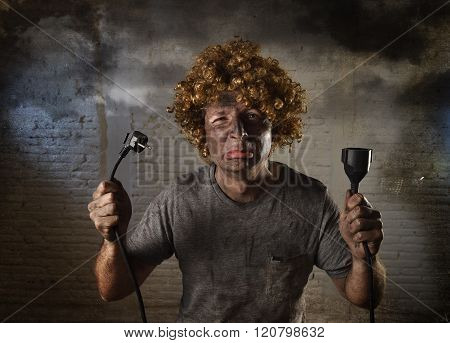 young man with funny curly wig holding electrical cable smoking after domestic accident with dirty burnt face and shock electrocuted expression in electricity DIY repairs danger concept in black smoke background