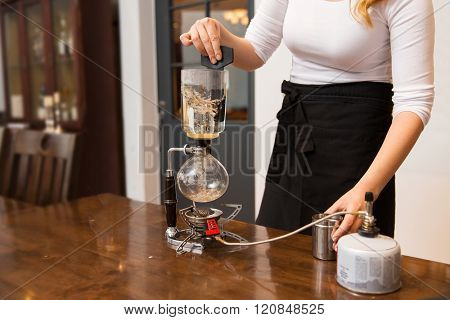 equipment, coffee shop, people and technology concept - close up of woman with butane gas burner heating water in siphon coffeemaker at cafe bar or restaurant kitchen