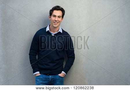 Happy man leaning on a grey wall. Handsome young man in casual leaning against a grey background and copy space. Young businessman looking at camera with hands in pocket.