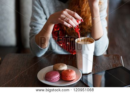 Young woman with red curly hair,and this spends time at a table in a café,stirs sugar in the white Cup of cappuccino coffee,red nail Polish,on the table is a white plate with multi colored French vanilla cookies