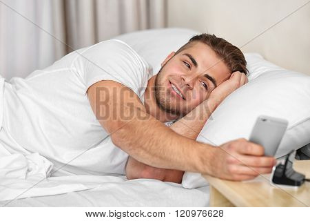 Beautiful young man waking up with mobile alarm clock
