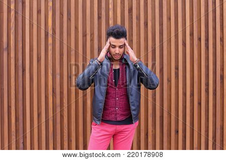 Handsome Young Arabic Male Man, Student Feels Headache, Touching Head, Trying to Ease Pain, Feels Discomfort, Standing on Background of Wooden Panel Stairs Outdoors. Confident Guy Arab Dressed in Black T-Shirt Over Maroon Shirt and Wears Black Leather Jac
