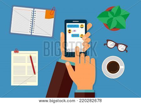 Hand holding smartphone with text messages on the screen. On the desktop, Notepad, pen, flower, coffee Cup and papers. The view from the top.