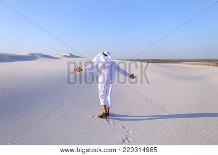 Stately young sheikh walks on foot along bottomless desert leaving traces on sand, enjoying warmth of day. Attractive Muslim smiles widely and spreads hands to sides, straightens clothes in wind and whirls around, delighted by breadth of desert. Swarthy, 