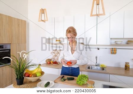 Attractive young woman cuts green cucumber for salad with sharp knife, takes red juicy tomato and several times throws up with smile on face, sniffs freshness of lettuce leaves and takes up shredding vegetables. Blonde at top of modern light kitchen