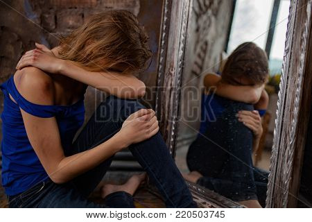 Beaten young woman victim of domestic violence and abuse cries and sits on floor near mirror with bruises and wounds on her body. Сoncept of domestic violence, sexual violence and cruelty.