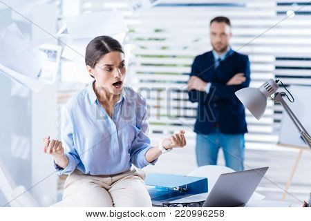 Furious woman. Emotional angry young woman sitting on the table and clenching her fists while shouting while her calm colleague standing behind her back with his arms crossed