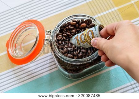 Woman Hand Putting A Roll One Hundred Dollar Bills Into Glass Jar With Coffee Beans On A Tablecloth.