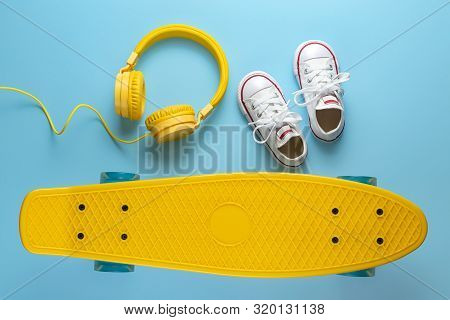 Yellow Headphones, White Sneakers And Skateboard Or Pennyboard On Blue Background. Music Concept.