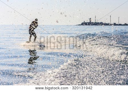 Wakeboarder Glides On Sea Surface Towed By Motor Boat With Splashes Scattering Around At Sunset. Wak
